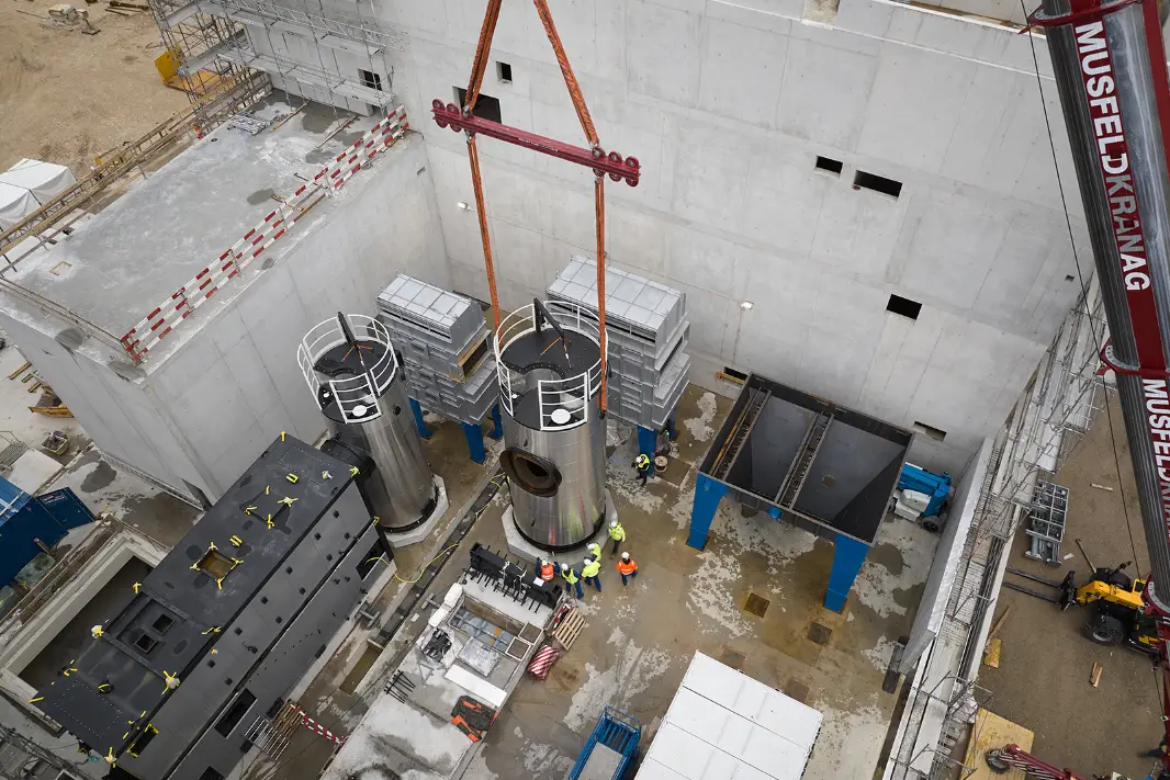 Chantier de la centrale énergétique UpTown Basel. De grands réservoirs cylindriques en métal sont soulevés en position par une grue sous la surveillance d'ouvriers.