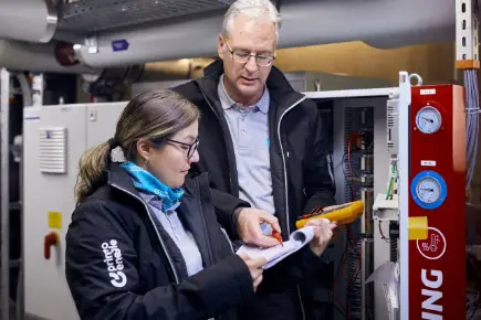 Two Primeo Energie technicians in work clothes and with tools stand in front of a control panel in an industrial plant. A woman is holding documents while the man points to something in the document.