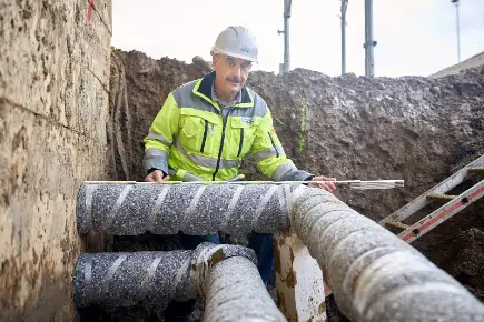Primeo Energie employee in yellow-grey safety clothing and hard hat working on a heat pipe. He is standing in a trench and holding a measuring device as he checks the large insulated pipes.