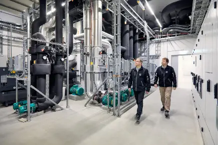 Two technicians from Primeo Energie walk through BaseLink's industrial heating centre with its large pipelines, valves and machines and check the technical equipment.