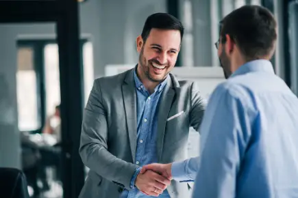 Two men in an office shake hands and smile. Windows and office furnishings can be seen in the background.