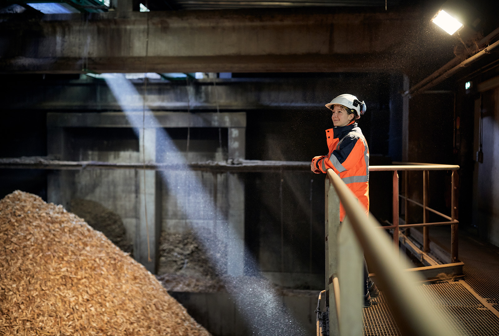 Une collaboratrice de Primeo, vêtue d'une tenue de travail et d'un casque de protection, se tient sur une passerelle métallique surélevée d'une installation de production de copeaux de bois et regarde un grand tas de copeaux de bois.