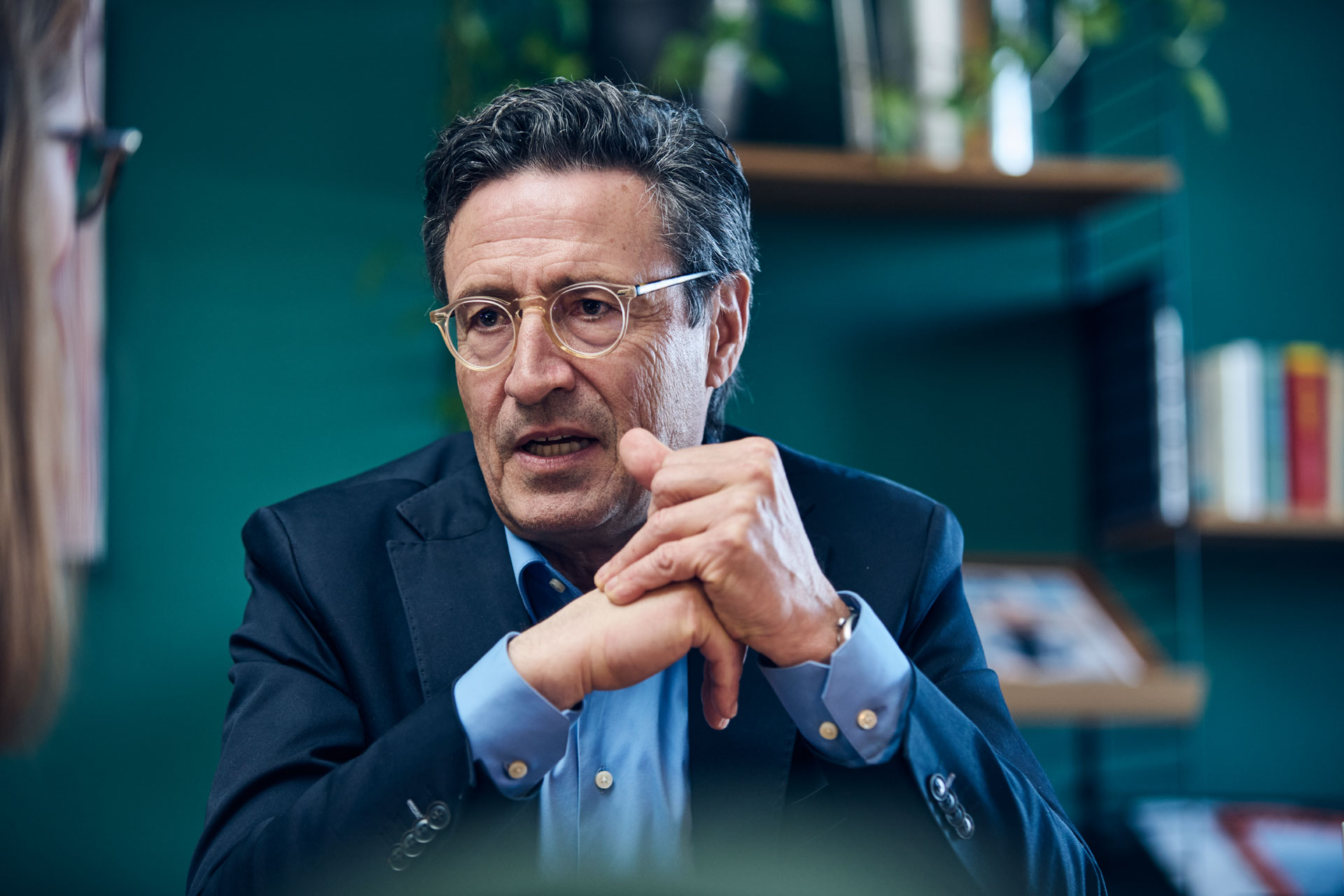 Ein Mann mittleren Alters mit Brille, dunklem Sakko und hellblauem Hemd spricht aufmerksam mit gefalteten Händen in einer modernen Büroumgebung.
