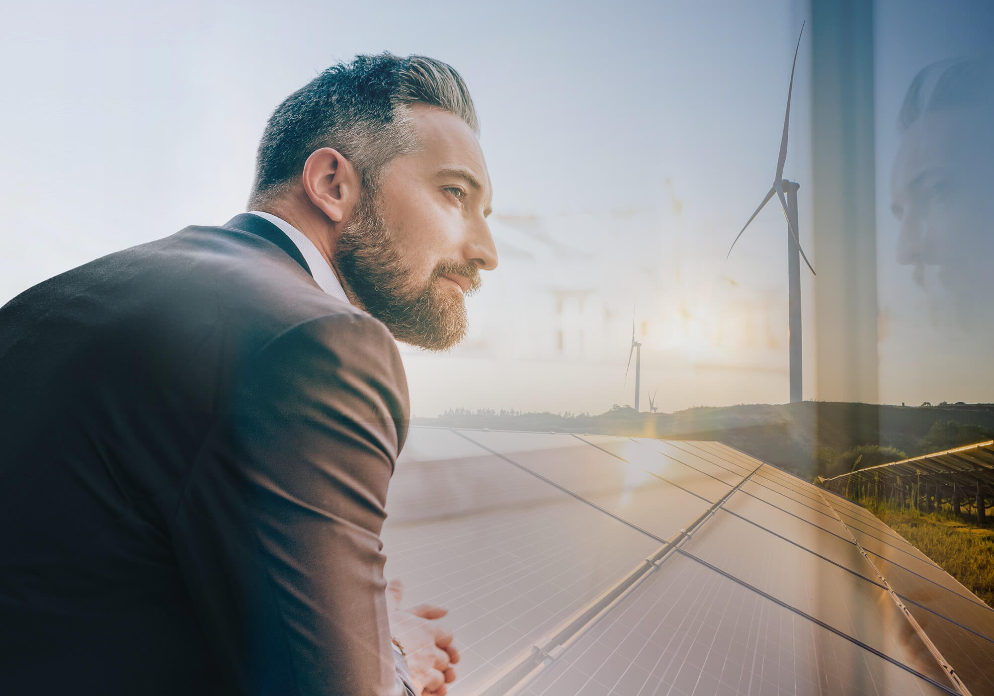 A thoughtful businessman in a suit looking through a window. In the background are superimposed images of solar panels and wind turbines at sunset.