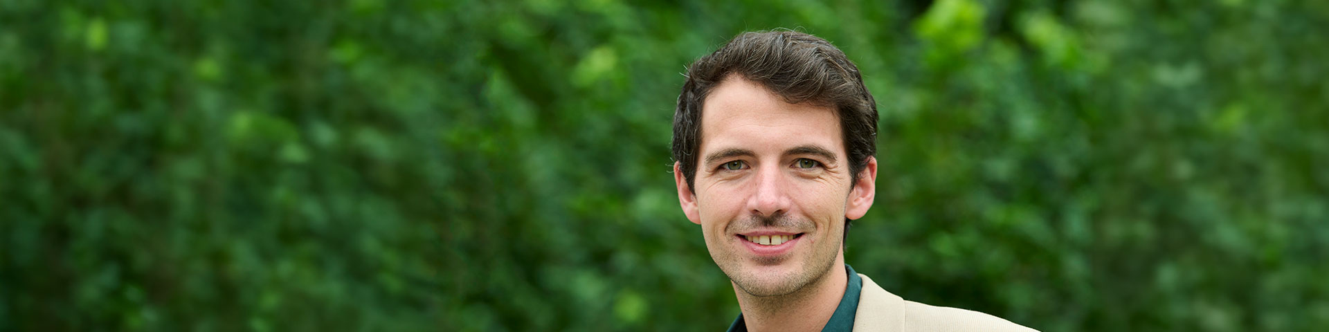Ein lächelnder junger Mann mit kurzen braunen Haaren und beigem Sakko vor einem grünen Blätterhintergrund.