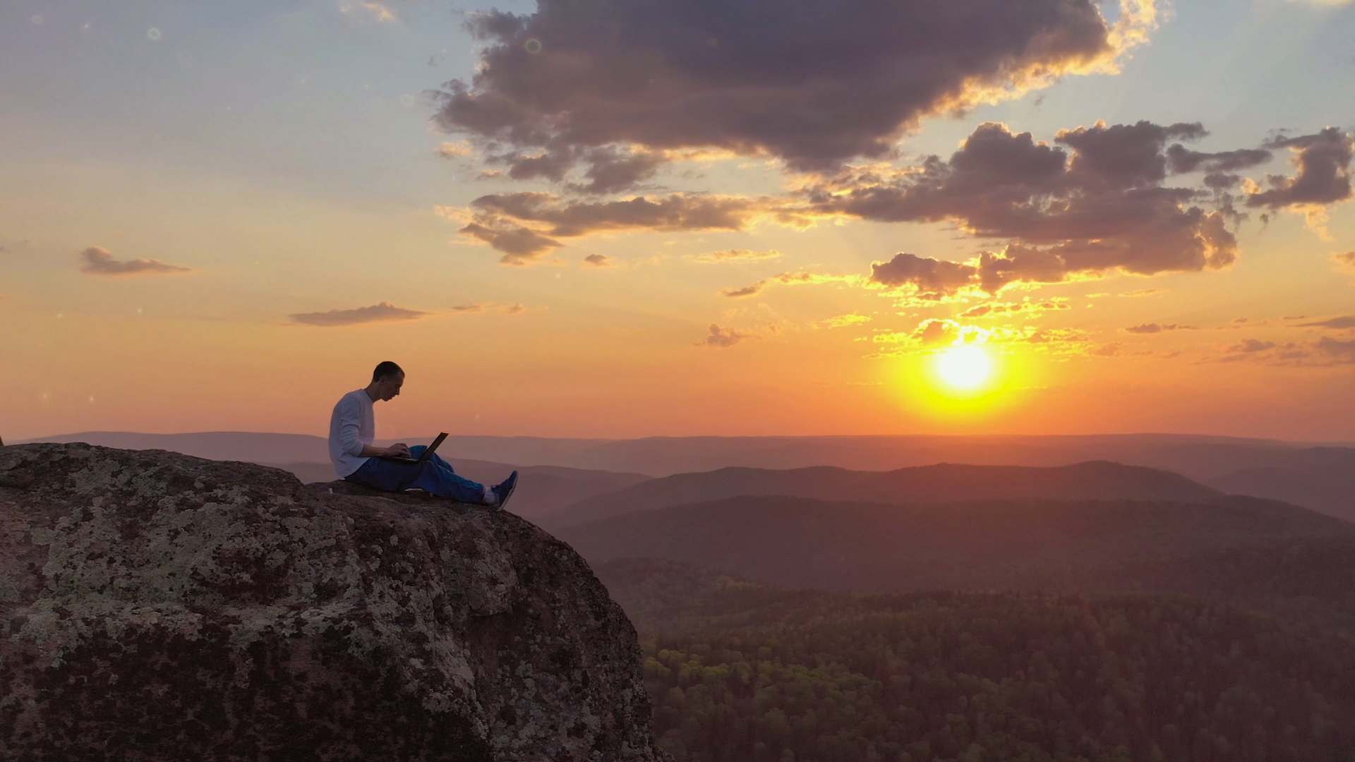 Homme assis sur un grand rocher au milieu d'un vaste paysage, travaillant sur un ordinateur portable au coucher du soleil.