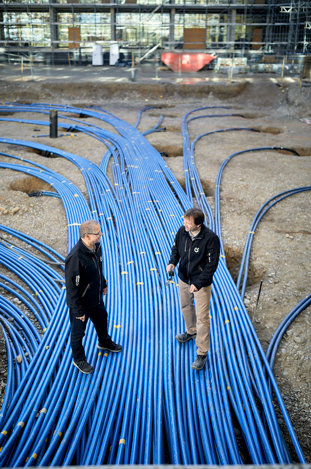 Deux techniciens de Primeo Energie se tiennent sur le chantier de BaseLink, entourés de nombreux tuyaux bleus enterrés qui courent sur le sol. En arrière-plan, on peut voir le bâtiment en construction avec ses échafaudages.