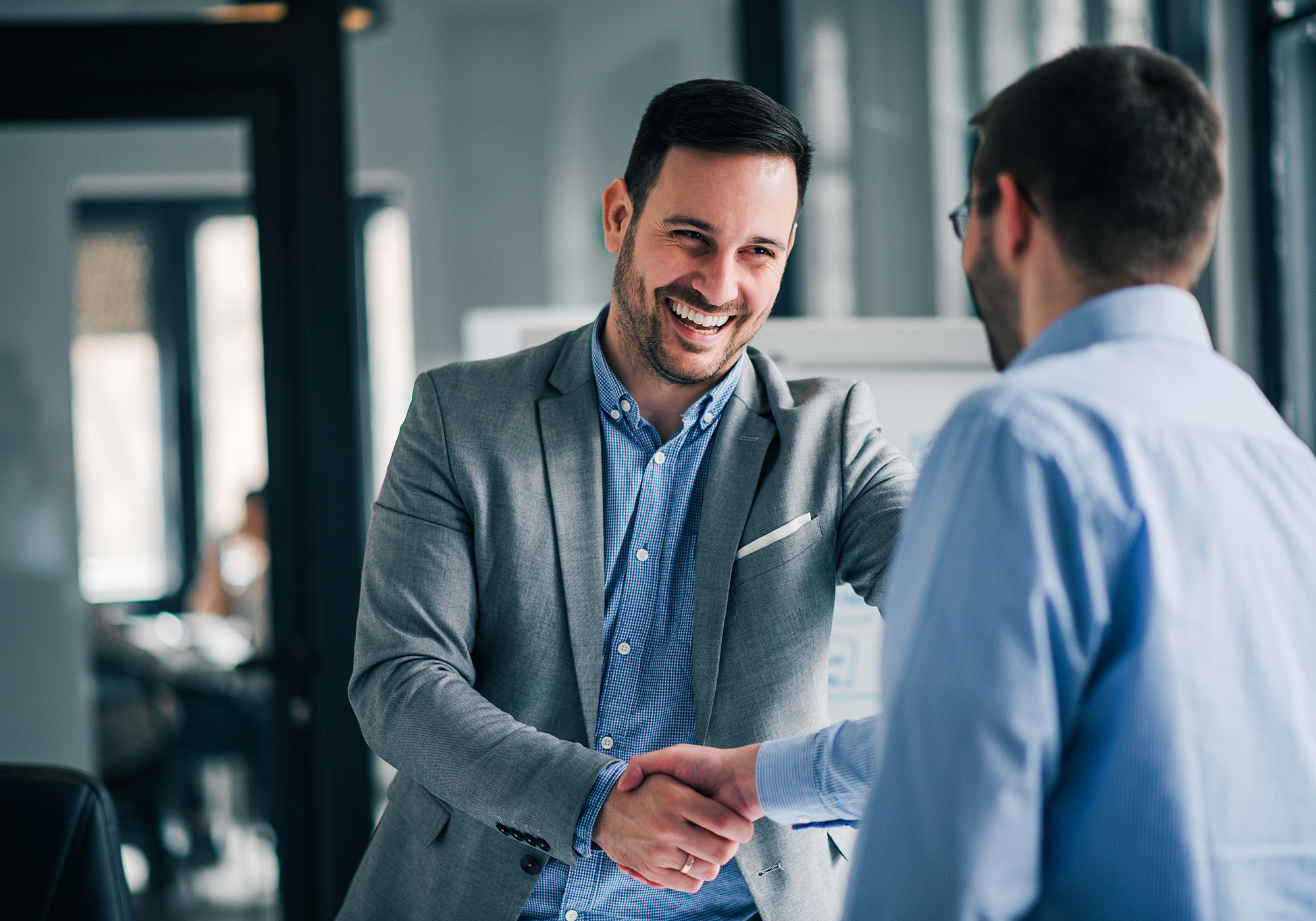 Two men in an office shake hands and smile. Windows and office furnishings can be seen in the background.