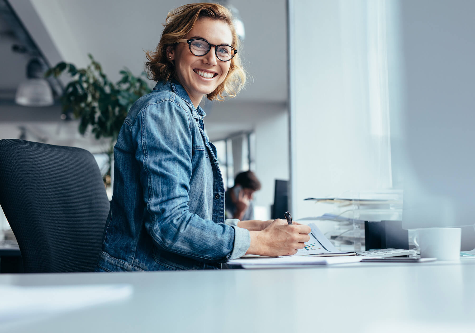 Une femme portant des lunettes et une veste en jean est assise à un bureau dans un bureau moderne et sourit.
