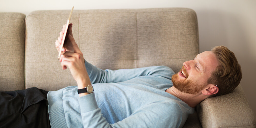 Un homme souriant est allongé sur un canapé, détendu, et tient une tablette dans ses mains en la regardant.